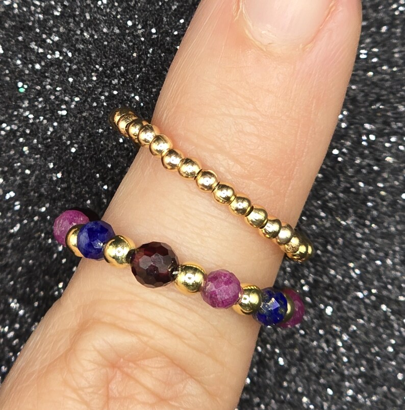 May include: Two rings on a finger. One is a gold beaded ring. The other ring has a gold band with alternating ruby, sapphire, and garnet gemstones. The rings are set against a sparkly black background.