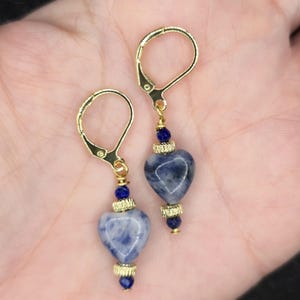 May include: A pair of heart-shaped earrings with gold-colored leverback closures. The earrings feature blue and white speckled heart-shaped stones, accented with gold-colored metal and small blue beads. The earrings are displayed against a neutral background.