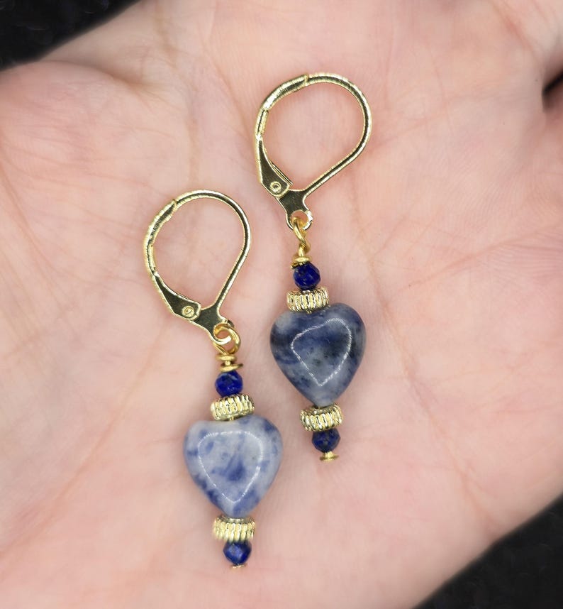 May include: A pair of heart-shaped earrings with gold-colored leverback closures. The earrings feature blue and white speckled heart-shaped stones, accented with gold-colored metal and small blue beads. The earrings are displayed against a neutral background.