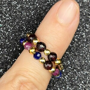 May include: Two beaded rings on a finger. One ring features dark red, gold, and purple beads. The other ring has dark red, gold, and blue beads. The rings are made of small, round beads strung together with gold-colored spacers.