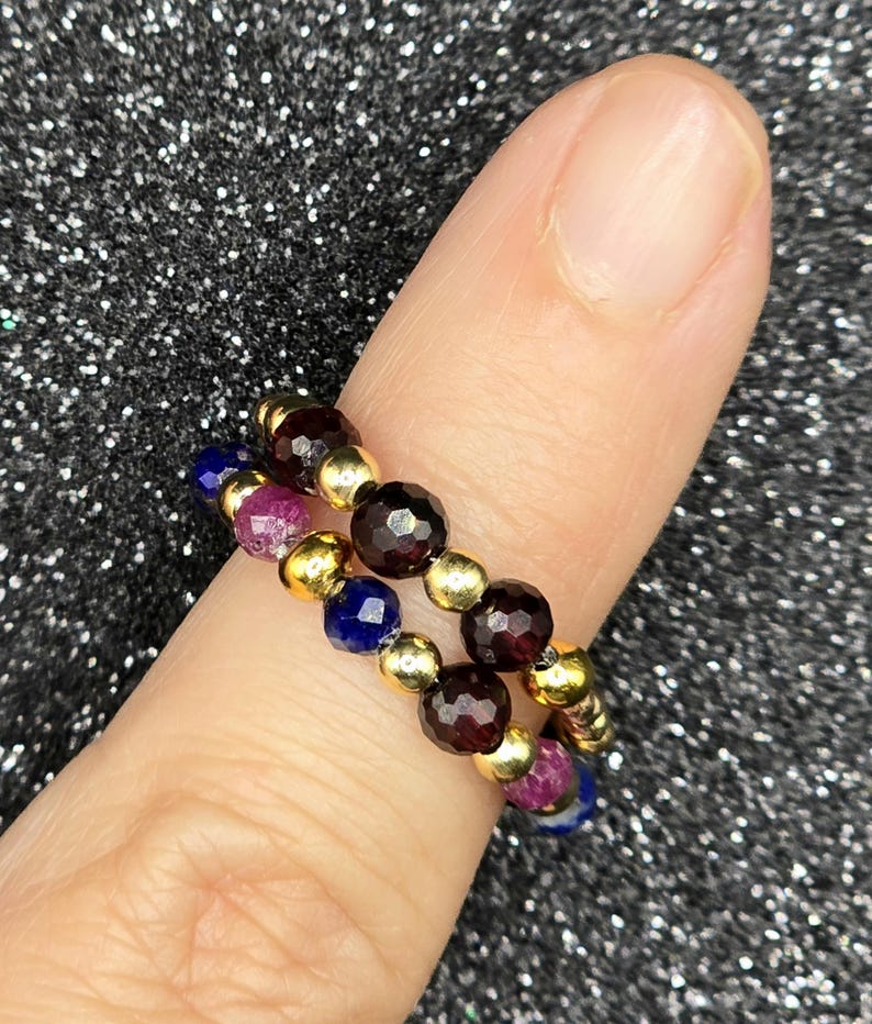 May include: Two beaded rings on a finger. One ring features dark red, gold, and purple beads. The other ring has dark red, gold, and blue beads. The rings are made of small, round beads strung together with gold-colored spacers.