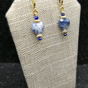 May include: A pair of gold-tone earrings with heart-shaped blue and white stone pendants. Each earring features a gold-tone lever back, a small blue bead, a gold-tone accent, and a larger heart-shaped stone pendant. The earrings are displayed on a textured gray surface.