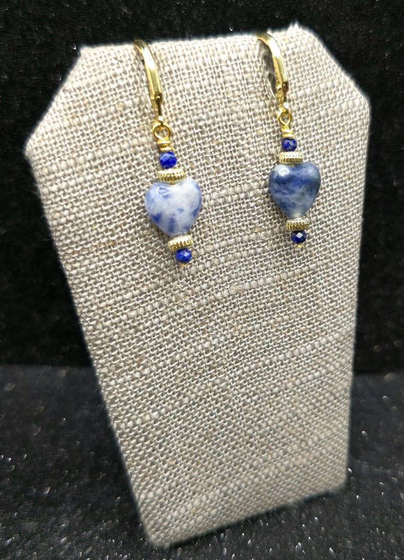 May include: A pair of gold-tone earrings with heart-shaped blue and white stone pendants. Each earring features a gold-tone lever back, a small blue bead, a gold-tone accent, and a larger heart-shaped stone pendant. The earrings are displayed on a textured gray surface.