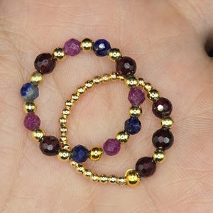 May include: Two beaded rings. One ring features alternating dark red, purple, and blue gemstones with gold beads. The other ring is made of small gold beads. Jewelry.