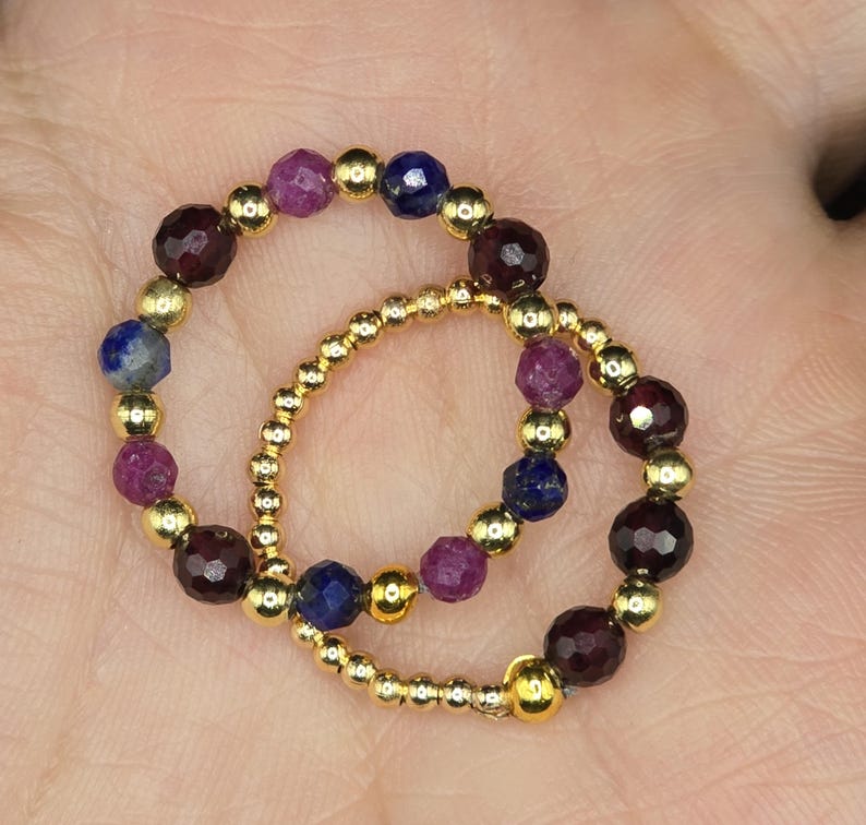 May include: Two beaded rings. One ring features alternating dark red, purple, and blue gemstones with gold beads. The other ring is made of small gold beads. Jewelry.