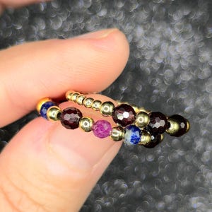 May include: Two beaded rings with gold-colored metal accents. The rings feature a variety of gemstones, including deep red, purple, and blue beads. The rings are held between fingers against a blurred background.