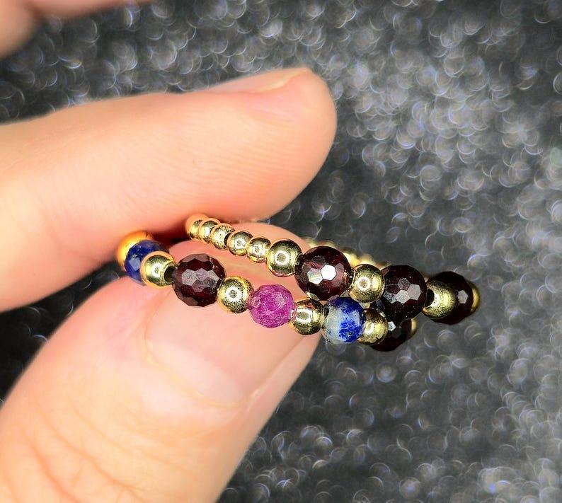 May include: Two beaded rings with gold-colored metal accents. The rings feature a variety of gemstones, including deep red, purple, and blue beads. The rings are held between fingers against a blurred background.