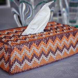 May include: A rectangular tissue box cover featuring a zig-zag pattern in orange, brown, and cream. The cover has the text "ellishdesigns" on the side. White tissues are visible, emerging from the top opening. The design is geometric.