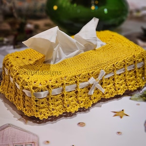 Pode incluir: Uma capa de caixa de lenços de crochê amarelo mostarda com detalhes em fita branca e um pequeno laço. A caixa retangular contém lenços brancos. O design apresenta crochet vazado e uma borda ondulada.