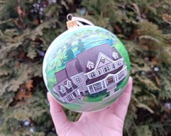Könnte beinhalten: Ein handbemalter Weihnachtsschmuck mit einer detaillierten Darstellung eines Hauses mit grauem Dach und weißem Zierrat. Der Schmuck hat einen grünen und blauen Hintergrund und eine goldfarbene Metallkappe mit weißem Band zum Aufhängen.