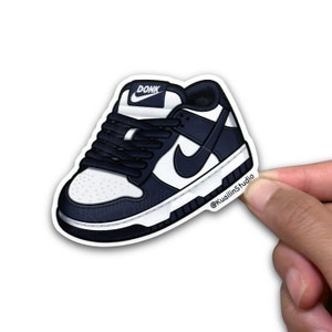 May include: A sticker featuring a navy blue and white sneaker illustration. The shoe has a white sole, a navy blue swoosh, and navy blue laces. The word "DONK" is printed on the tongue of the shoe. The sticker is held by a hand.