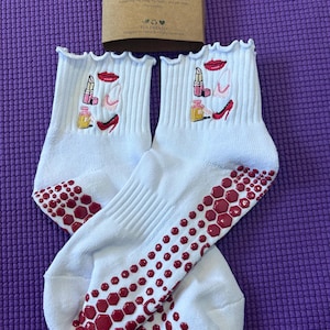 Puede incluir: Un par de calcetines tobilleros blancos con la parte superior con volantes y suelas de agarre rojas. Cada calcetín presenta diseños de maquillaje bordados, incluyendo lápiz labial, un frasco de perfume y un tacón alto. Se ve una tarjeta marrón con texto.