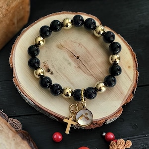 Könnte beinhalten: Ein Perlenarmband mit abwechselnd dunkelblauen, gesprenkelten Perlen und polierten goldfarbenen Metallkugeln. Das Armband enthält zwei hängende Anhänger: ein schlichtes goldfarbenes Kreuz und einen runden Anhänger mit einer Berggrafik.
