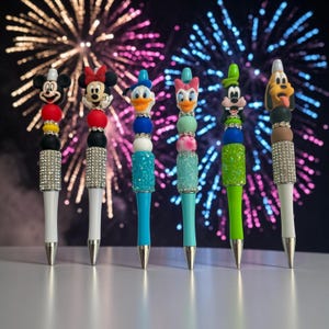 May include: Six pens featuring character toppers including Mickey Mouse, Minnie Mouse, Donald Duck, Daisy Duck, Goofy, and Pluto. Each pen has colorful beads, rhinestone accents, and a glittery barrel section, standing upright in a row.
