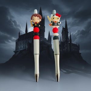 Könnte beinhalten: Zwei silberfarbene Stifte mit dekorativen Aufsätzen. Der linke Stift hat eine braunhaarige Figuren-Applikation mit Brille und Schal. Der rechte Stift zeigt eine weiße Katzenfigur mit roter Schleife, Brille und Schal. Beide haben rote und schwarze Perlen.