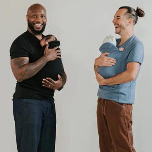 May include: Two people holding babies in baby carriers. One person wears a black t-shirt and dark blue jeans, the other wears a blue t-shirt and brown trousers. The baby carriers are black and blue.