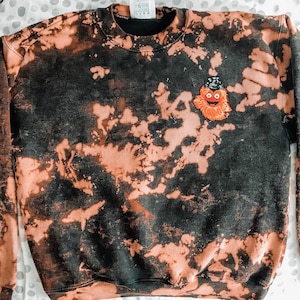 Könnte beinhalten: Ein schwarzes und orangefarbenes Batik-Sweatshirt mit einer kleinen, orangefarbenen und schwarzen Stickfigur auf der Vorderseite. Die Figur trägt einen schwarzen Hut und hat ein breites Lächeln.