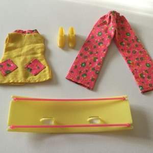 May include: A vintage doll clothing set featuring a yellow dress with pink floral patches, pink floral pants, yellow shoes, and a yellow bed frame with pink accents. The set is likely from the 1970s or 1980s.