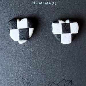 Puede incluir: Un par de pendientes en forma de corazón a cuadros en blanco y negro. Los pendientes están hechos a mano y tienen una superficie texturizada.
