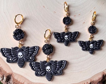 May include: Three pairs of black and gold dangle earrings featuring a black moth with a white skull design and black glitter accents.