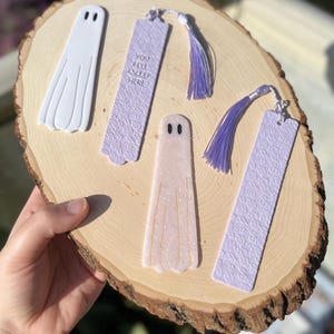 Könnte beinhalten: Vier handgefertigte Lesezeichen mit Geistermotiven. Zwei Lesezeichen sind weiß mit einer Geister-Silhouette, eines ist lavendelfarben mit einer Geister-Silhouette und Glitzer, und eines ist lavendelfarben mit dem Text "You fell asleep here".
