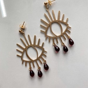 May include: A pair of gold eye-shaped earrings with dangling burgundy teardrop beads.