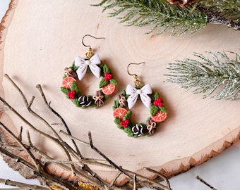 May include: A pair of gold dangle earrings featuring a green wreath with red berries, orange slices, cinnamon sticks, and a white bow. The earrings are on a wooden surface.