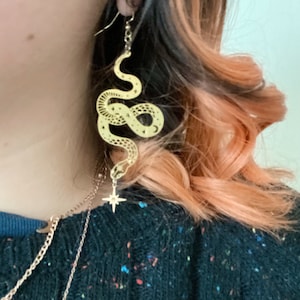 Op de afbeelding: Gouden slangen oorbellen met een sterbedel die aan de onderkant hangt. De slang is versierd met een sterrenpatroon.