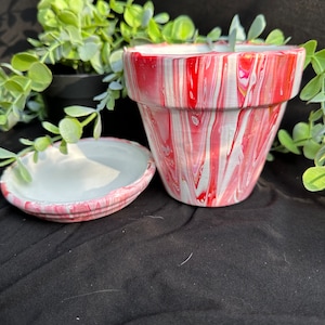 May include: A white and red swirl painted terracotta planter with a matching saucer. The planter has a wide mouth and a rounded bottom.