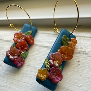 May include: A pair of teal blue hoop earrings with a floral design. The earrings feature a cluster of pink, orange, and yellow flowers with green leaves.