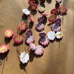 May include: A collection of handmade seashell earrings in various styles and colors. The earrings feature pink, yellow, and purple hues, with some designs incorporating gold hoops and stud posts. The earrings are arranged on a brown surface.
