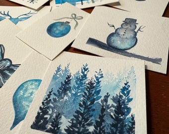 May include: A set of ten watercolor paintings on white cardstock, each depicting a different winter-themed image. The images include a snowflake, a plaid pattern, a Christmas ornament, a snowman, a forest, a deer, and a few abstract designs.