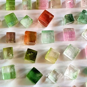 May include: A collection of 36 small, faceted, cubic gemstones in various shades of green, pink, red, and clear. The gemstones are arranged in a grid pattern on a white surface.