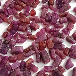 Może przedstawiać: Duża kolekcja małych, surowych, czerwonych i fioletowych rubinów. Rubiny mają różne kształty i rozmiary, a niektóre mają lekko spiczasty koniec.