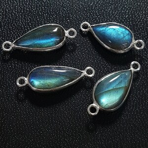 May include: Four teardrop-shaped labradorite pendants with silver-colored metal settings. The stones display iridescent blue and green hues. Each pendant has a loop at both ends for attaching to a chain or other jewelry component. The pendants are arranged on a textured black surface.