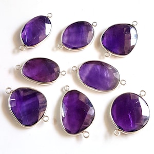 Op de afbeelding: Acht amethist hangers met zilverkleurige connectoren. Elke hanger heeft een vrije ovale vorm met een glad, gepolijst oppervlak.