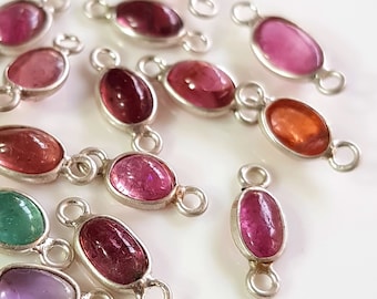 Puede incluir: Una colección de conectores de piedras preciosas ovaladas en varios colores, incluyendo rosa, rojo, verde y morado. Cada piedra preciosa está engastada en un marco de metal plateado con bucles en cada extremo para la fijación. Las piedras están sobre una superficie blanca.