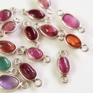 Puede incluir: Una colección de conectores de piedras preciosas ovaladas en varios colores, incluyendo rosa, rojo, verde y morado. Cada piedra preciosa está engastada en un marco de metal plateado con bucles en cada extremo para la fijación. Las piedras están sobre una superficie blanca.