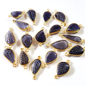 Puede incluir: Una colección de dijes de hojas de piedras preciosas azul oscuro con bordes y bucles de metal dorado para la fabricación de joyas. Algunas hojas tienen detalles grabados. Los dijes están dispersos sobre una superficie blanca.