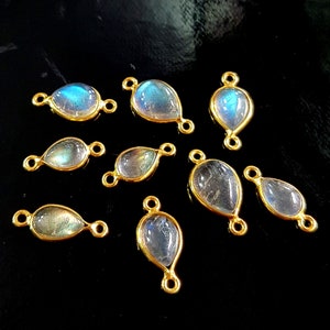 May include: Nine teardrop-shaped labradorite gemstone pendants with gold-colored bezels. The iridescent stones display blue and green hues. Each pendant has small loops on either end for attaching to jewelry. The pendants are displayed on a black surface.