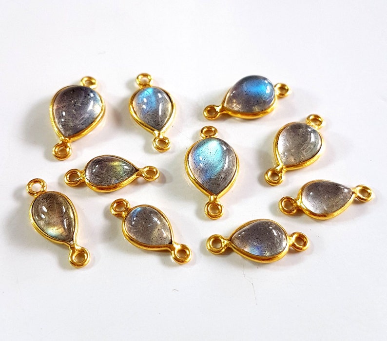 May include: A collection of teardrop-shaped labradorite gemstone pendants with gold-colored metal settings. Each pendant has a loop at the top and bottom for attaching to a chain or other jewelry component. The gemstones display iridescent blue and gray hues.