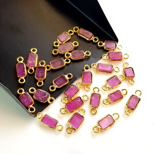 Op de afbeelding: Een verzameling rechthoekige, magenta edelsteenconnectoren met goudkleurige metalen frames. De edelstenen hebben een enigszins ruwe textuur. De connectoren zijn gerangschikt op een wit oppervlak met een zwarte lade.