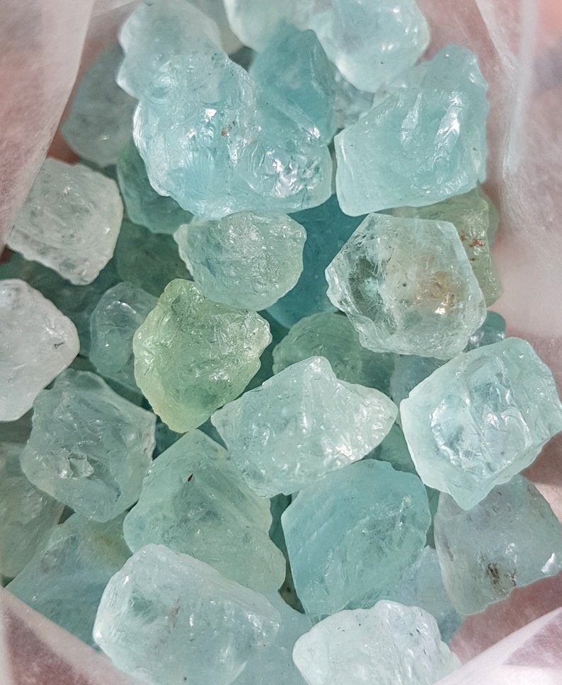 Puede incluir: Una vista cercana de una bolsa llena de cristales de aguamarina en bruto. Los cristales son de varios tonos de azul y verde, y algunos tienen un ligero brillo.
