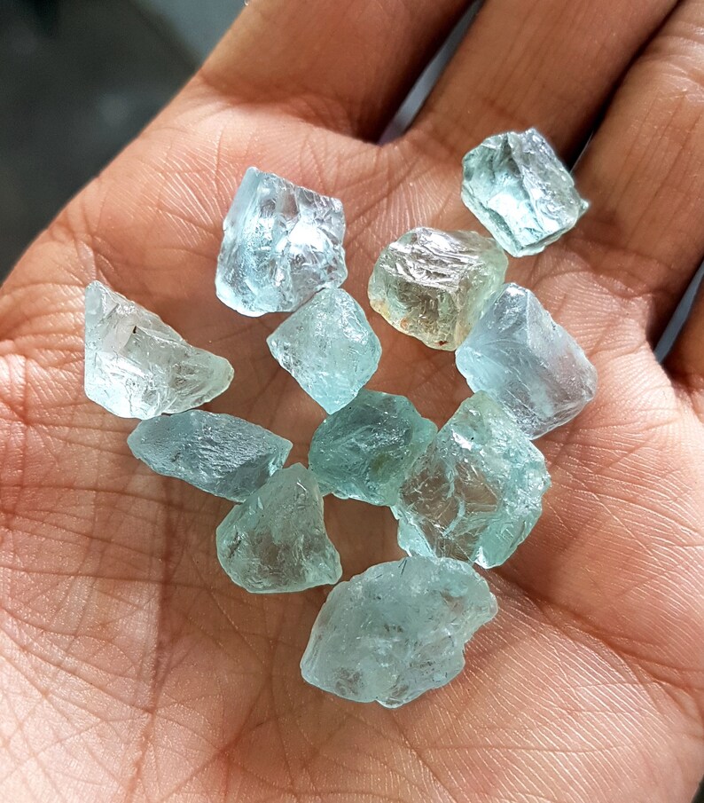 Puede incluir: Una colecci&oacute;n de piedras preciosas de aguamarina azul claro, en bruto y sin cortar, con varias formas y tama&ntilde;os. Las piedras son transl&uacute;cidas y tienen un ligero tono verde.