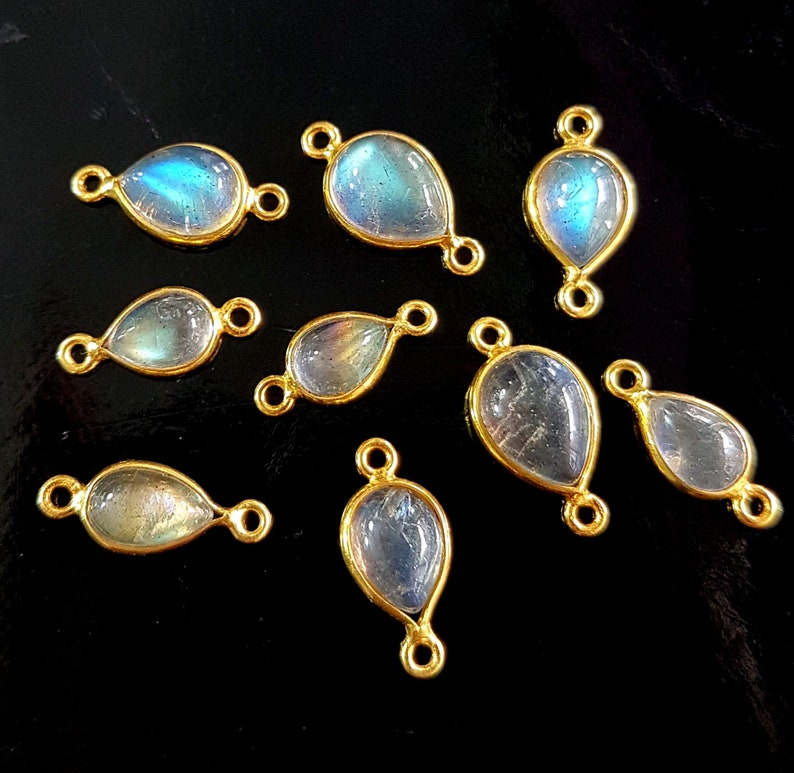 May include: A collection of teardrop-shaped labradorite gemstone pendants with gold-colored bezels and connecting loops. The iridescent stones display blue and green hues. The pendants are arranged on a black surface, showcasing their polished finish.