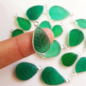 Pode incluir: Pingentes de pedras preciosas verdes em forma de folha com acabamento em metal prateado. Os pingentes têm um design de folha esculpida e são em forma de gota. A imagem mostra vários pingentes, um deles segurado por um dedo.