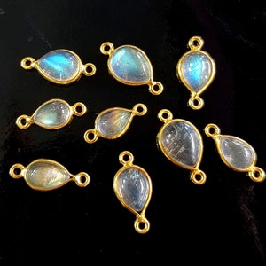 May include: A collection of teardrop-shaped labradorite gemstone pendants with gold-colored bezels and connecting loops. The iridescent stones display blue and green hues. The pendants are arranged on a black surface, showcasing their polished finish.