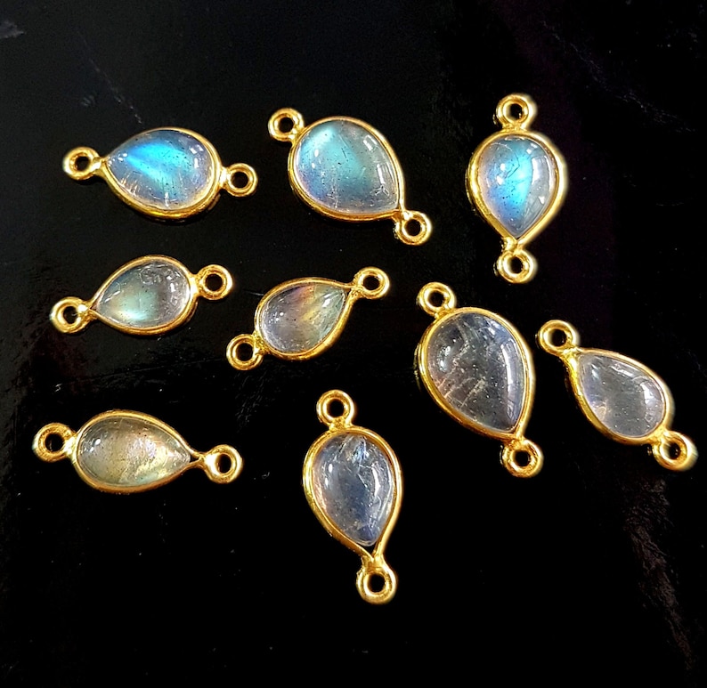May include: A collection of teardrop-shaped labradorite gemstone pendants with gold-colored bezels and connecting loops. The iridescent stones display blue and green hues. The pendants are arranged on a black surface, showcasing their polished finish.