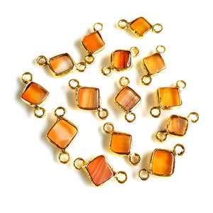 Può includere: Una collezione di connettori in metallo color oro, ciascuno con una gemma arancione di forma quadrata o a diamante. Le gemme hanno un aspetto naturale e striato. Ogni connettore ha due anelli per il fissaggio.