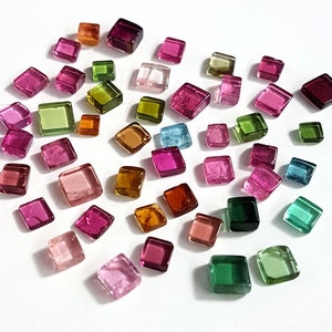Puede incluir: Una colección de pequeñas piedras preciosas de corte cuadrado en varios colores, incluyendo rosa, verde, naranja y azul. Las gemas están dispersas sobre una superficie blanca, mostrando su calidad translúcida y bordes facetados. Las piedras preciosas son probablemente para la fabricación de joyas.
